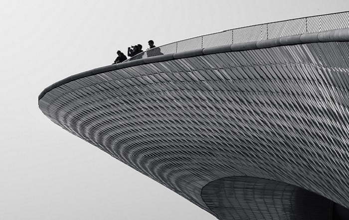 Black-and-white image of a large, curved architectural structure with people standing on top, showcasing its sweeping, futuristic design and textured surface